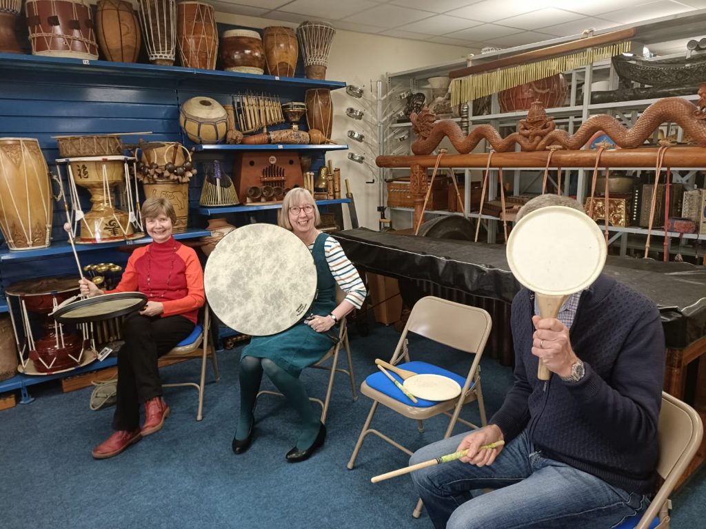 Image Description: A set of five photographs, showing Evelyn and members of the Volunteer team. All of the photos show a variety of the Volunteer team smiling, some with people sitting and playing Uchiwa Daikos and Remo Paddle Drums, and some photos with people stood casually chatting with a light buffet on a table in the middle of the room.